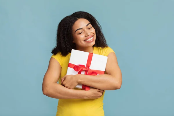 En iyi hediye. Neşeli Afro-Amerikalı kadın hediye kutusuna sarılıyor mavi stüdyo arka planında poz veriyor, gözleri kapalı gülümsüyor. — Stok fotoğraf