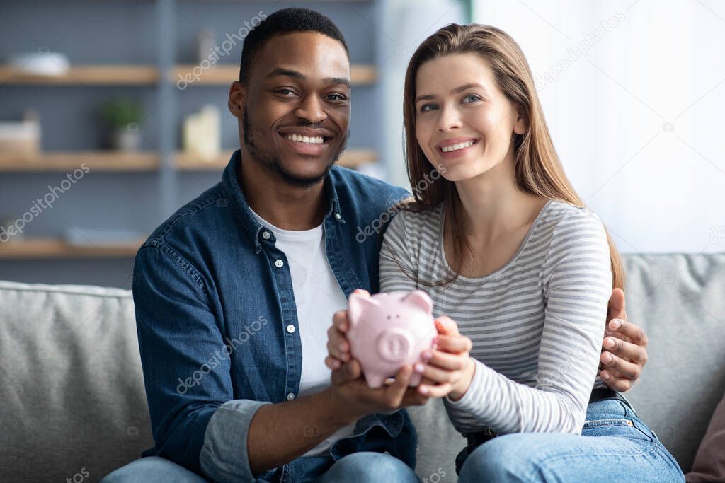 Happy Young Interracial Couple Holding Pink Piggybank And Smiling At Camera