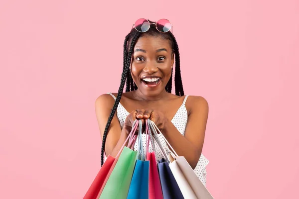 Encantadora joven mujer negra con racimos afro sosteniendo un montón de bolsas de compras en el fondo del estudio rosa —  Fotos de Stock