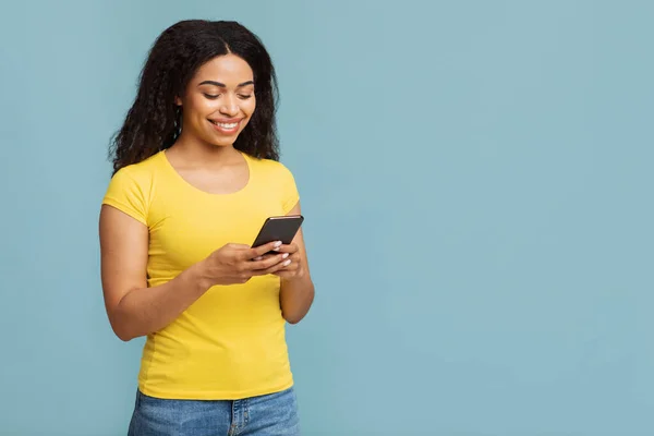 Alat trendi yang keren. Wanita afrika yang bersemangat menggunakan smartphone, berdiri di atas latar belakang biru dengan ruang fotokopi — Stok Foto