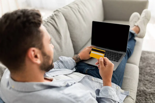 Vásárlásmánia és e-kereskedelem. Fiatal srác kezében hitelkártya, online vásárlás laptopon, ül a kanapén otthon — Stock Fotó