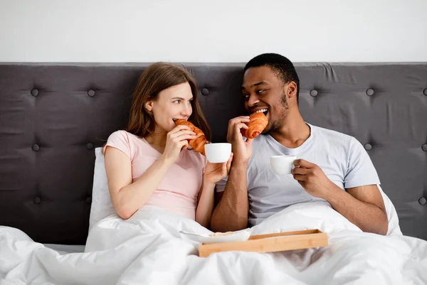 Affettuosa coppia multirazziale che fa colazione a letto, mangiare croissant freschi growth caffè a casa — Foto Stock