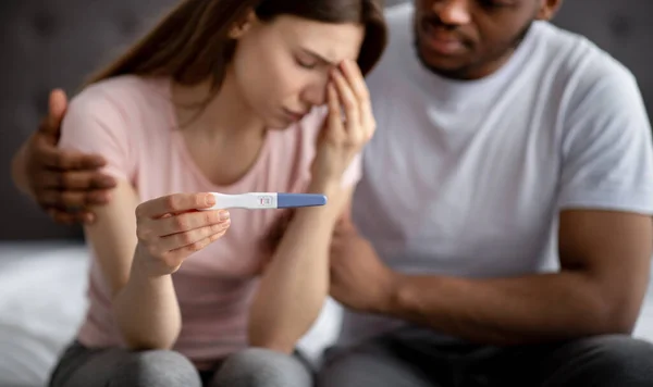 Verärgerte junge Frau mit negativem Schwangerschaftstest, ihr schwarzer Ehemann unterstützt sie im Schlafzimmer, selektiver Fokus — Stockfoto