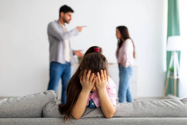 Семейный кризис. Маленькая девочка плачет, закрывая лицо руками, пока ее злые родители дерутся на заднем плане — стоковое фото
