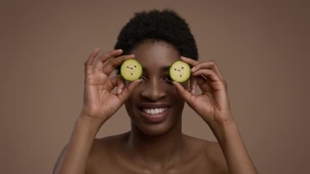 Mujer africana posando con rodajas de pepino cubriendo los ojos, fondo beige — Vídeos de Stock