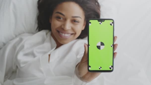 Gran aplicación móvil. Mujer afroamericana positiva mostrando teléfono inteligente con pantalla de croma verde, acostada en la cama — Vídeos de Stock