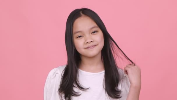 Маленькая кокетка. Милая игривая азиатская девушка играет с волосами и улыбается в камеру, флиртует и улыбается, розовый фон — стоковое видео