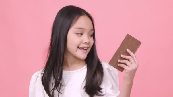 Tatlı sevgilim. Küçük tatlı Asyalı kız çikolata tutuyor ve gülüyor, büyük tatlı yiyor, pembe stüdyo geçmişi var. — Stok video