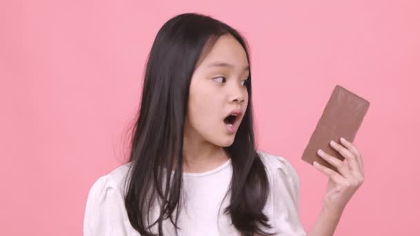 Dulce diente. Estudio retrato de pequeña chica asiática disfrutando de gran barra de chocolate y postre mordaz, rosa fondo del estudio — Vídeos de Stock