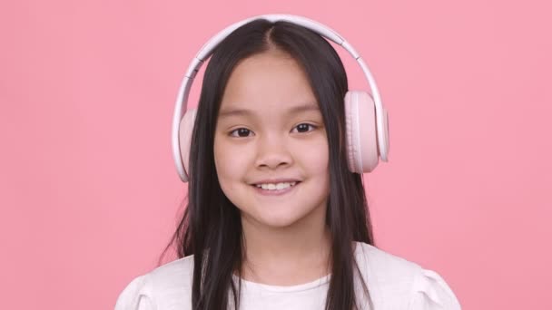 Modern zenei technológiák. Imádnivaló kis ázsiai lány visel nagy vezeték nélküli headset mosolygó kamera — Stock videók