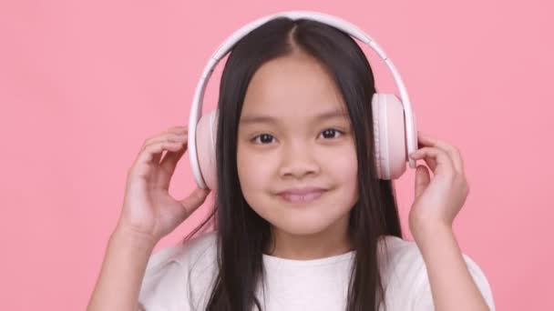 Linda chica asiática en auriculares inalámbricos escuchando música dinámica, bailando y sonriendo a la cámara — Vídeo de stock