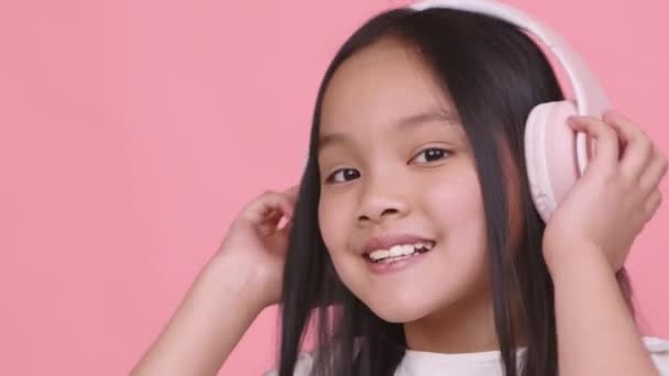 Muziekliefhebber. Vrolijk klein aziatisch meisje luisteren naar muziek in grote roze draadloze hoofdtelefoon, glimlachen naar de camera — Stockvideo