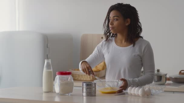 Acogedora cocina de la mañana. Joven mujer afroamericana pacífica mezclando huevos con batidor y mirando a un lado en la cocina — Vídeos de Stock