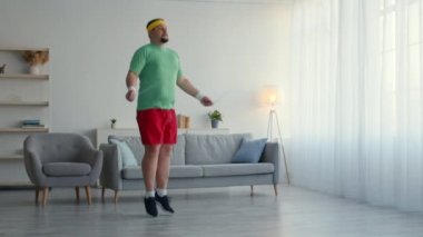 Spor kıyafetleri giyen tombul adam ip atlayıp evde zıplıyor spor antrenmanlarıyla kilo vermeye çalışıyor.