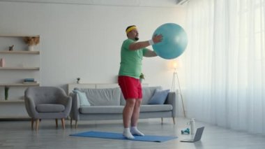 Yeni başlayanlar için online spor eğitimi. Tombul adam formda topla egzersiz yapıyor, hamleler yapıyor, dizüstü bilgisayarda özel ders izliyor.