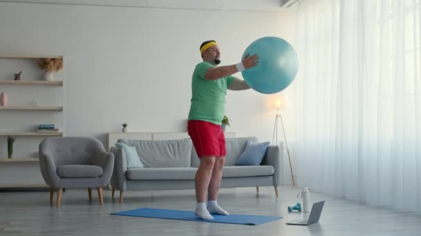 Online sport training for beginners. Chubby man exercising with fit ball, doing lunges, watching tutorial on laptop — Stockvideo