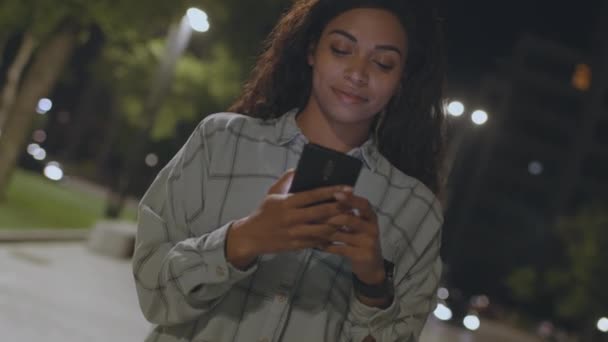 Tirada giratoria de la joven afroamericana enfocada establecimiento de redes en el teléfono inteligente en el parque de la noche y sonriendo — Vídeos de Stock