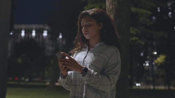 Online communication. Young african american lady chatting on smartphone with friends, standing in dark evening park — Stock video