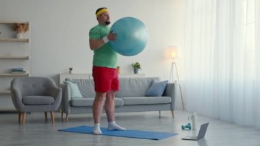 Aşırı kilolu adam internette egzersiz yapıyor, dizüstü bilgisayarda video dersi izliyor ve evde top oynarken çömelme egzersizleri yapıyor.