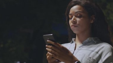 İnternet iletişimi. Ciddi bir Afro-Amerikan kadını karanlık akşam parkında yürüyor ve akıllı telefondan mesajlar okuyor.