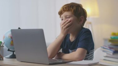 Evde eğitim çok yorucu. Sevimli küçük kızıl çocuk yorgun hissediyor, bilgisayardan online dersler izlerken esniyor.
