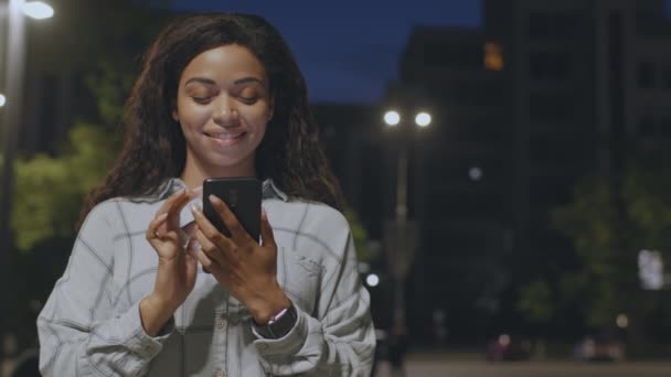 Urban lifestyle concept. Young positive african american woman texting on smartphone, standing in night park, free space — Stock video