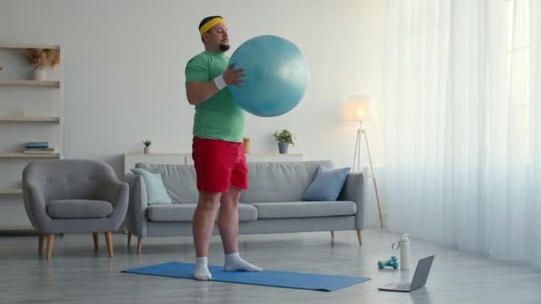 Übergewichtiger Mann trainiert online, schaut Video-Tutorial auf Laptop und übt tiefe Kniebeugen mit fittem Ball zu Hause — Stockvideo