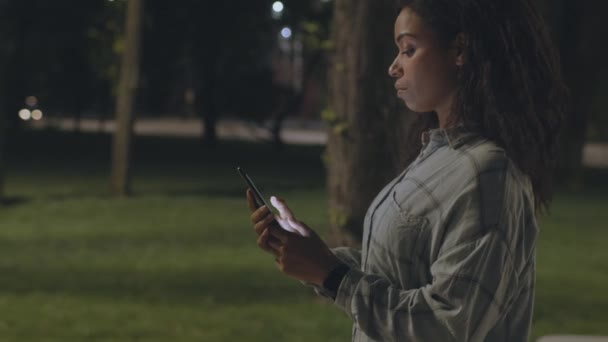 Stile di vita moderno e dipendenza gadget. Giovane donna afro-americana in rete su smartphone, passeggiando nel parco serale — Video Stock