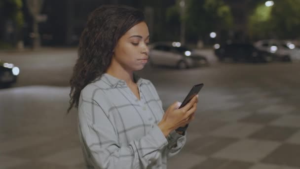 Peligro de la adicción a Internet. Joven afroamericana enfocada en leer noticias sobre teléfonos inteligentes cruzando la carretera a pie en la noche — Vídeo de stock