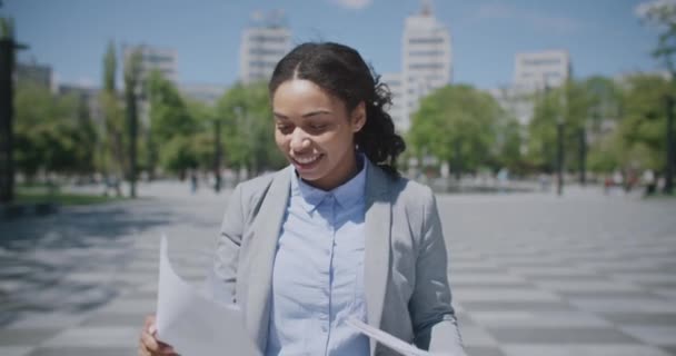 Afro-Amerikan kadın şehir parkında yürüyor, iş belgelerini okuyor ve gülümsüyor, iyi sonuçların tadını çıkarıyor. — Stok video