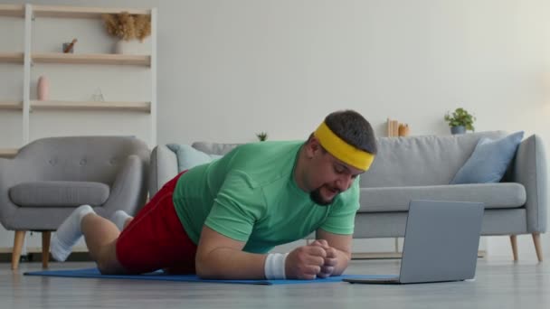 Chubby man exercising at home, greeting coach via video call on laptop and doing plank, exercising in living room — Stock Video