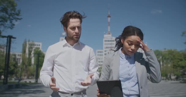 Problema de negócios. Multiétnico homem e mulher colegas de trabalho discutindo erro no projeto, andando ao ar livre com tablet digital — Vídeo de Stock