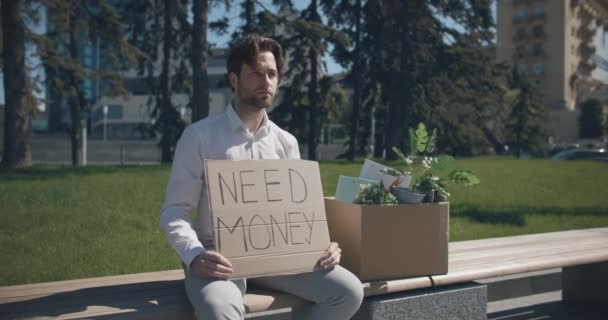 Увеличить портрет безработного молодого офисного работника, сидящего на скамейке с личными вещами, с плакатом "Нужные деньги" — стоковое видео