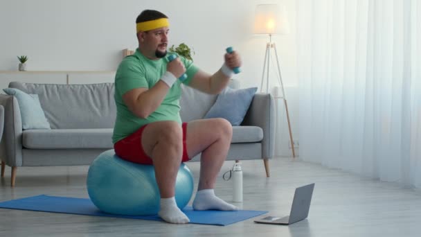 Hombre con sobrepeso sentado en la pelota en forma y haciendo ejercicio con pesas en casa, viendo clases de fitness en el portátil — Vídeo de stock