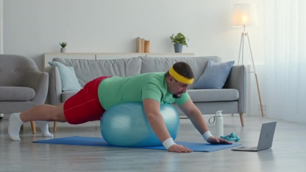 Pilates für Dicke. Übergewichtiger Mann liegt auf Fitball und schaut Fitnessstunde am Laptop, macht Dehnübungen — Stockvideo