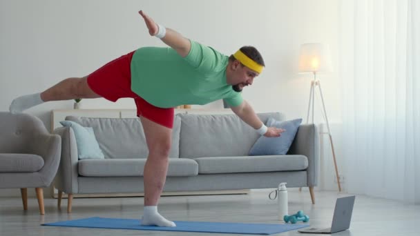 Funny chubby man in sportive outfit, practicing balance is swallow position, watching online yoga lesson on laptop — Stockvideo