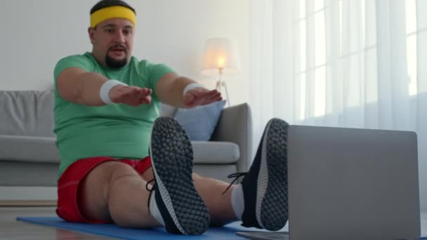 Músculos abdominales estirándose. Hombre regordete en ropa brillante entrenamiento en casa, ejercitando contra el ordenador portátil en el suelo — Vídeo de stock