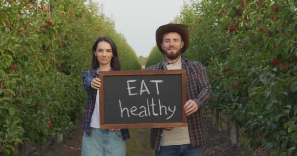 Valores familiares. Pareja joven de horticultores posando entre su huerto de manzanas y mostrando jabalí con texto Come sano — Vídeos de Stock