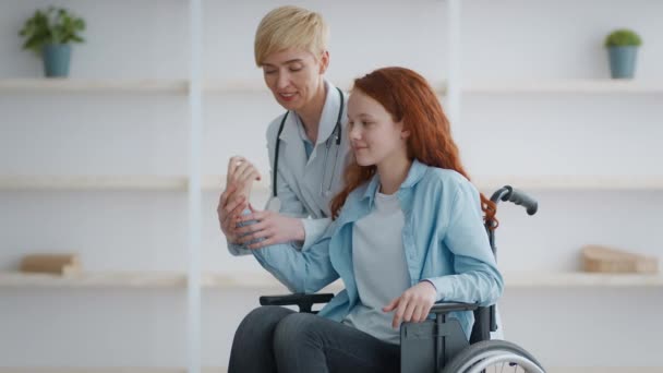 Femme médecin professionnel réchauffant les poignets de petite rousse paralysée assise en fauteuil roulant à la maison, au ralenti — Video