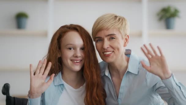 Petite rousse handicapée assise en fauteuil roulant, embrassant sa mère et agitant la main à la caméra à la maison — Video