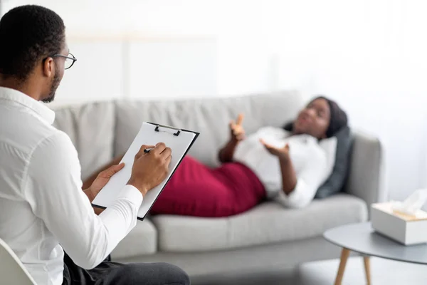 Молода чорна жінка за консультацією з психологом, лежачи на дивані в офісі, пояснює свою проблему доктору — стокове фото