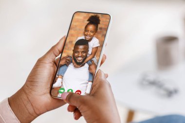 Siyahi kadın, neşeli kocası ve kızıyla sohbet ederken, akıllı telefonu kullanarak evinden ailesiyle konuşuyor.