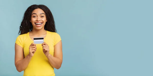 Mujer afroamericana llena de alegría sosteniendo tarjeta de crédito, fondo azul, panorama con espacio para copiar — Foto de Stock