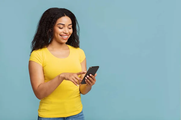 Aplikasi keren. Happy african american woman using smartphone, standing over blue studio background with free space — Stok Foto