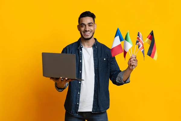 Cheerful arab guy holding laptop and international flags on yellow — Stock fotografie