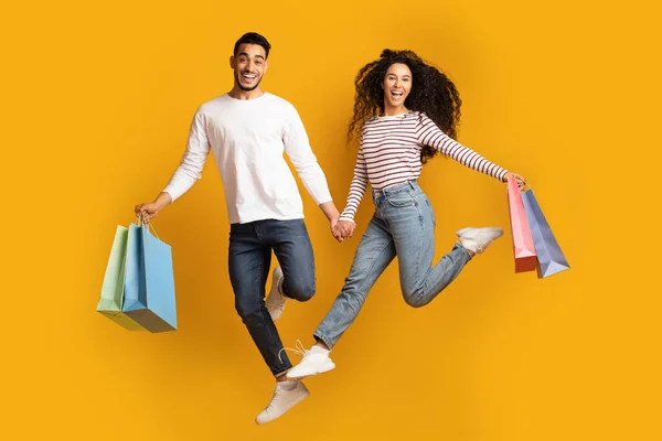 Alegre pareja árabe sosteniendo las manos y saltando con bolsas de compras —  Fotos de Stock