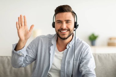 İnternetteki öğretmenlerin portresi. Kulaklık takan mutlu yakışıklı adam web kamerasına el sallıyor, selam veriyor.