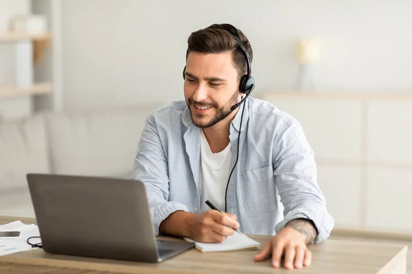 Nuevos conocimientos en línea en casa. Hombre joven en auriculares tomando notas y mirando el ordenador portátil en la sala de estar — Foto de Stock