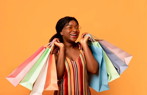 Cliente satisfecho. Joven mujer negra alegre en ropa de verano sosteniendo bolsas de compras brillantes, posando y mirando a un lado —  Fotos de Stock