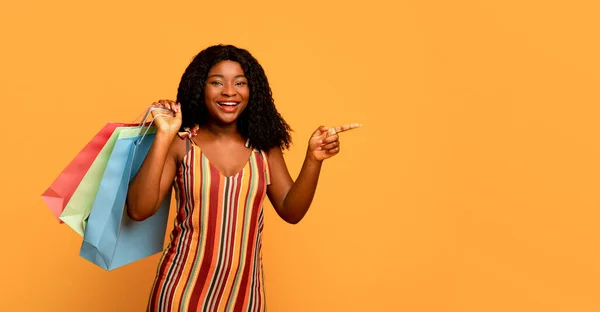 Grandes vendas. Retrato de jovem mulher negra feliz com sacos de compras apontando para o espaço em branco sobre fundo laranja — Fotografia de Stock